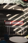 Sunlight streams through overhead beams, casting shadows on a wall featuring a prominent sign with bold red letters stating 'GOOD BUSINESS'. The environment appears to be indoors with modern architectural elements and minimalistic decor. Potted plants and furniture create a relaxed, professional atmosphere.