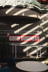 Sunlight streams through overhead beams, casting shadows on a wall featuring a prominent sign with bold red letters stating 'GOOD BUSINESS'. The environment appears to be indoors with modern architectural elements and minimalistic decor. Potted plants and furniture create a relaxed, professional atmosphere.