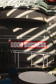 Sunlight streams through overhead beams, casting shadows on a wall featuring a prominent sign with bold red letters stating 'GOOD BUSINESS'. The environment appears to be indoors with modern architectural elements and minimalistic decor. Potted plants and furniture create a relaxed, professional atmosphere.
