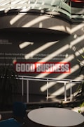 Sunlight streams through overhead beams, casting shadows on a wall featuring a prominent sign with bold red letters stating 'GOOD BUSINESS'. The environment appears to be indoors with modern architectural elements and minimalistic decor. Potted plants and furniture create a relaxed, professional atmosphere.