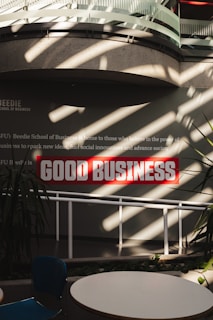 Sunlight streams through overhead beams, casting shadows on a wall featuring a prominent sign with bold red letters stating 'GOOD BUSINESS'. The environment appears to be indoors with modern architectural elements and minimalistic decor. Potted plants and furniture create a relaxed, professional atmosphere.
