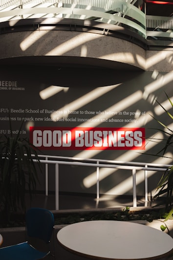 Sunlight streams through overhead beams, casting shadows on a wall featuring a prominent sign with bold red letters stating 'GOOD BUSINESS'. The environment appears to be indoors with modern architectural elements and minimalistic decor. Potted plants and furniture create a relaxed, professional atmosphere.