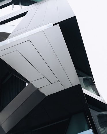 A modern architectural structure with angular, geometric shapes and clean lines. The surface appears smooth and metallic, reflecting light subtly. The design is minimalist, highlighting the precision of the construction.