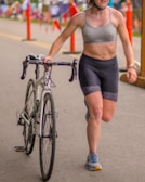 A person wearing a grey sports bra and black cycling shorts is walking alongside a bicycle on an outdoor road. They are wearing a helmet, and in the background, there are orange traffic cones and spectators on a sunny day.