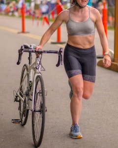 A person wearing a grey sports bra and black cycling shorts is walking alongside a bicycle on an outdoor road. They are wearing a helmet, and in the background, there are orange traffic cones and spectators on a sunny day.