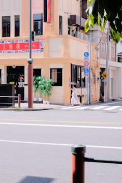 A street corner with a pale yellow building that features a variety of national flags above the entrance. There are two large potted plants by the front door. A person with an umbrella is walking across the crosswalk in front of the building. Various street signs, poles, and traffic symbols are visible, along with a pedestrian crossing sign.