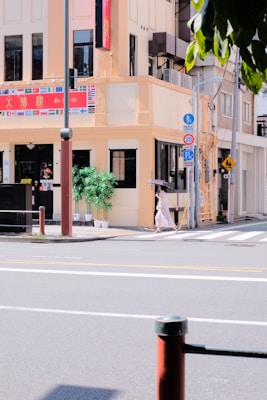 A street corner with a pale yellow building that features a variety of national flags above the entrance. There are two large potted plants by the front door. A person with an umbrella is walking across the crosswalk in front of the building. Various street signs, poles, and traffic symbols are visible, along with a pedestrian crossing sign.