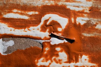 An abstract image featuring a blend of warm and earthy tones. The scene includes prominent rusty orange brushstrokes across a rough textured surface, with a piece of curled metal embedded in the foreground. Variations in color and texture create a dynamic, fluid composition.