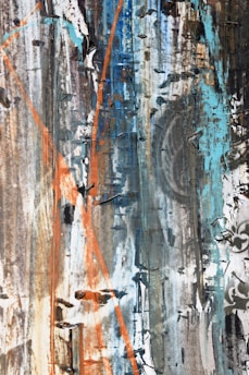 A textured abstract artwork featuring a mix of colors including blue, orange, and gray. The surface appears weathered and layered, with paint peeling and streaks running vertically. There are hints of geometric shapes and faded patterns throughout.