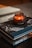 A rich mahogany-colored candle sits on top of two stacked books, one of which is titled 'AMAZING ITALY'. The scene exudes a warm and cozy atmosphere, with soft lighting highlighting the textures and colors of the objects.