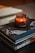 A rich mahogany-colored candle sits on top of two stacked books, one of which is titled 'AMAZING ITALY'. The scene exudes a warm and cozy atmosphere, with soft lighting highlighting the textures and colors of the objects.