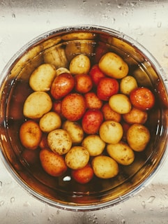 A collection of small, round potatoes in varying shades of yellow and red are submerged in a metal bowl filled with water. The bowl is situated on a stainless steel surface, which is wet with droplets of water.
