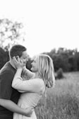 A couple stands closely together in an open field, gazing into each other's eyes with affectionate expressions. The woman gently holds the man's face with both hands, while the man wraps his arms around her waist. The background features blurred trees and tall grass, creating a serene and intimate atmosphere.
