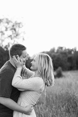 A couple stands closely together in an open field, gazing into each other's eyes with affectionate expressions. The woman gently holds the man's face with both hands, while the man wraps his arms around her waist. The background features blurred trees and tall grass, creating a serene and intimate atmosphere.