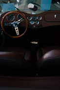The interior of a vintage car featuring a wooden dashboard with multiple circular dials and gauges. A classic steering wheel with a distinctive design is prominent, and the leather seats are visible in the foreground. There are blue papers with the number 7978 visible through the windshield.