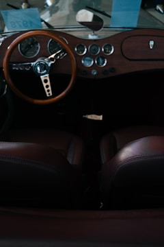 The interior of a vintage car featuring a wooden dashboard with multiple circular dials and gauges. A classic steering wheel with a distinctive design is prominent, and the leather seats are visible in the foreground. There are blue papers with the number 7978 visible through the windshield.
