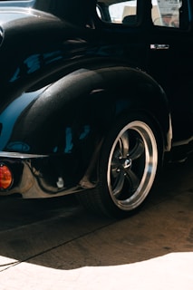 The photograph features the rear end of a sleek black vintage car with smooth, glossy curves. The chrome trim on the car and the polished surface reflect light beautifully, highlighting the intricate design of the vehicle. The metallic rim of the wheel, along with the tire, is prominently displayed. The car is positioned on a light brown concrete surface with visible textures.