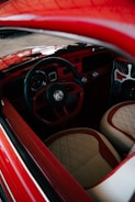 A vintage car interior with a retro-styled steering wheel and dashboard. The seats have a quilted pattern with red and beige colors, and the door panels are detailed with artistic accents.
