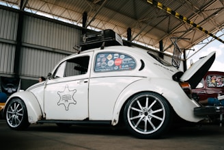 A vintage white Volkswagen Beetle with custom modifications is parked inside a large warehouse structure. The car features multiple stickers on the side windows, a roof rack, and stylish alloy wheels. The engine hood is open, revealing part of the rear-engine compartment. The surroundings include a high ceiling with beams and a partially visible red car.