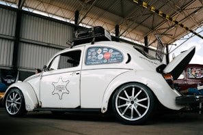 A vintage white Volkswagen Beetle with custom modifications is parked inside a large warehouse structure. The car features multiple stickers on the side windows, a roof rack, and stylish alloy wheels. The engine hood is open, revealing part of the rear-engine compartment. The surroundings include a high ceiling with beams and a partially visible red car.