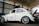 A vintage white Volkswagen Beetle with custom modifications is parked inside a large warehouse structure. The car features multiple stickers on the side windows, a roof rack, and stylish alloy wheels. The engine hood is open, revealing part of the rear-engine compartment. The surroundings include a high ceiling with beams and a partially visible red car.