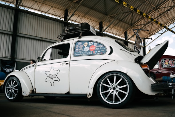 A vintage white Volkswagen Beetle with custom modifications is parked inside a large warehouse structure. The car features multiple stickers on the side windows, a roof rack, and stylish alloy wheels. The engine hood is open, revealing part of the rear-engine compartment. The surroundings include a high ceiling with beams and a partially visible red car.