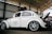 A vintage white Volkswagen Beetle with custom modifications is parked inside a large warehouse structure. The car features multiple stickers on the side windows, a roof rack, and stylish alloy wheels. The engine hood is open, revealing part of the rear-engine compartment. The surroundings include a high ceiling with beams and a partially visible red car.