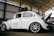 A vintage white Volkswagen Beetle with custom modifications is parked inside a large warehouse structure. The car features multiple stickers on the side windows, a roof rack, and stylish alloy wheels. The engine hood is open, revealing part of the rear-engine compartment. The surroundings include a high ceiling with beams and a partially visible red car.