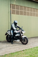 A person in full motorcycle riding gear, including a helmet, is seated on a stationary sport motorcycle. The motorcycle is white with distinctive design elements and is parked on a concrete surface adjacent to a green wall with a grid-like pattern. There is a patch of grass alongside the concrete area.