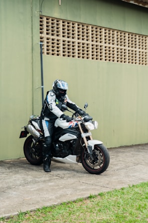 A person in full motorcycle riding gear, including a helmet, is seated on a stationary sport motorcycle. The motorcycle is white with distinctive design elements and is parked on a concrete surface adjacent to a green wall with a grid-like pattern. There is a patch of grass alongside the concrete area.