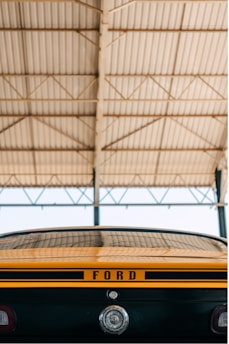 The image features the rear view of a classic car under a large metal roof with a clear sky visible in the background. The car has a stripe design with the word 'FORD' prominently displayed on the trunk and a circular emblem below it.