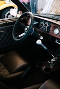 A vintage car interior featuring a classic steering wheel, wooden dashboard, and leather seats. The manual gear shift is prominent, and the dashboard includes analog dials and gauges. Outside the car, part of a yellow vehicle with retro styling is visible.