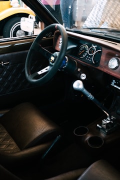 A vintage car interior featuring a classic steering wheel, wooden dashboard, and leather seats. The manual gear shift is prominent, and the dashboard includes analog dials and gauges. Outside the car, part of a yellow vehicle with retro styling is visible.