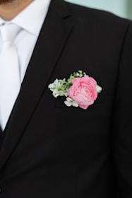 A man wearing a black suit jacket with a white shirt and tie. A pink rose boutonniere with white and green accents is placed in the suit's lapel pocket.