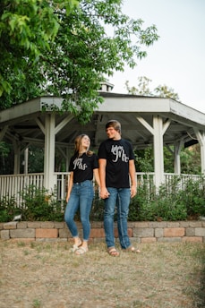 A couple stands in front of a white gazebo, holding hands and smiling at each other. They wear matching black t-shirts with white text and jeans. Surrounding them are lush green trees and plants, and the ground is covered with short grass and a stone border.