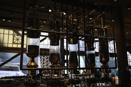 Intricate coffee brewing equipment with several large, transparent vessels filled with coffee beans, connected by shiny metal pipes. This setup is situated in a well-lit interior space with large windows. The industrial design and warm lighting create a sophisticated ambiance.