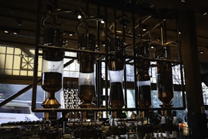 Intricate coffee brewing equipment with several large, transparent vessels filled with coffee beans, connected by shiny metal pipes. This setup is situated in a well-lit interior space with large windows. The industrial design and warm lighting create a sophisticated ambiance.