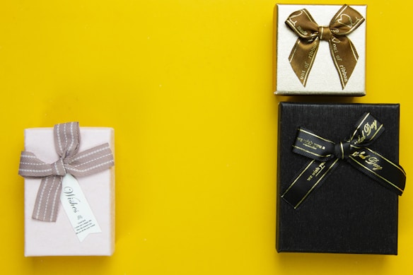 Three gift boxes are arranged on a bright yellow background. The first box is wrapped in pale paper with a gray ribbon and a small tag. The second box is smaller, wrapped in gold paper with a brown ribbon. The third box is black with a black ribbon featuring gold text.