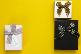 Three gift boxes are arranged on a bright yellow background. The first box is wrapped in pale paper with a gray ribbon and a small tag. The second box is smaller, wrapped in gold paper with a brown ribbon. The third box is black with a black ribbon featuring gold text.
