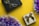 Two gift boxes are positioned against a vibrant yellow background, with one box being black with a black ribbon and the other gold with a brown ribbon. Purple and white tinsel adds a festive touch to the image.