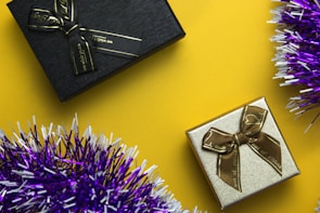 Two gift boxes are positioned against a vibrant yellow background, with one box being black with a black ribbon and the other gold with a brown ribbon. Purple and white tinsel adds a festive touch to the image.