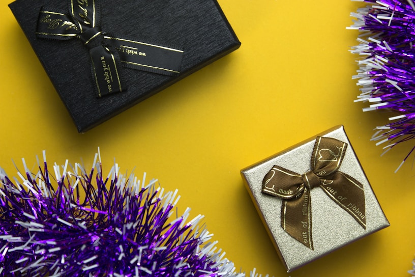 Two gift boxes are positioned against a vibrant yellow background, with one box being black with a black ribbon and the other gold with a brown ribbon. Purple and white tinsel adds a festive touch to the image.