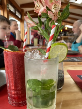 Two refreshing drinks with striped straws are placed on a table with pink flowers in the background. One drink appears to be a mojito with mint leaves, ice, and a lime slice, while the other is a red smoothie. Several people can be seen in a blurred background, suggesting a lively dining environment.