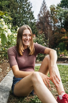 A person is sitting outdoors in a garden filled with various plants and flowers. They are smiling, wearing a brown short-sleeved top and denim shorts, and appear relaxed. The background showcases green foliage, white and yellow flowers, and tall trees.