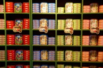 A set of neatly organized shelves displaying a variety of colorful canned seafood. The cans are arranged by type and color, featuring bright red, blue, and green labels. Some cans have decorative illustrations and tags with pricing, showcasing a vibrant and appealing presentation.
