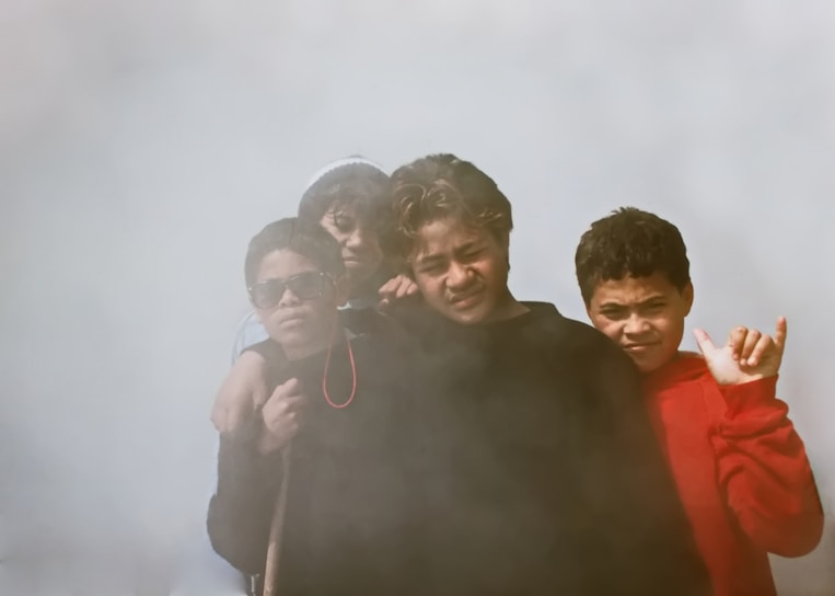 A group of four kids stand together, surrounded by a misty or foggy atmosphere. One child on the left wears sunglasses and dark clothing, while another on the right is wearing a bright red sweater and making a playful gesture. The expressions are relaxed, suggesting a casual group photo.