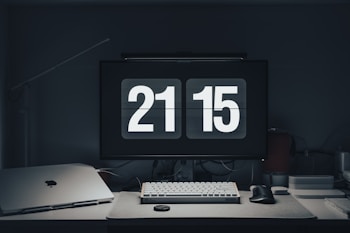 A modern workspace features a digital clock displaying 21:15 on a computer monitor. The desk is organized with a keyboard, mouse, laptop, and other tech accessories. The overall setting is dimly lit, creating a calm and focused atmosphere.