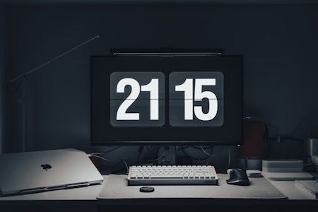 A modern workspace features a digital clock displaying 21:15 on a computer monitor. The desk is organized with a keyboard, mouse, laptop, and other tech accessories. The overall setting is dimly lit, creating a calm and focused atmosphere.