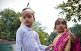 Two children are dressed in traditional attire, the boy in a white kurta with a headband and red markings on his forehead, while the girl is wearing a pink and gold sari, accessorized with jewelry including a forehead ornament. They are outdoors with trees in the background.
