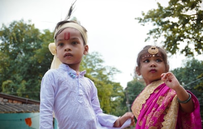 Two children are dressed in traditional attire, the boy in a white kurta with a headband and red markings on his forehead, while the girl is wearing a pink and gold sari, accessorized with jewelry including a forehead ornament. They are outdoors with trees in the background.